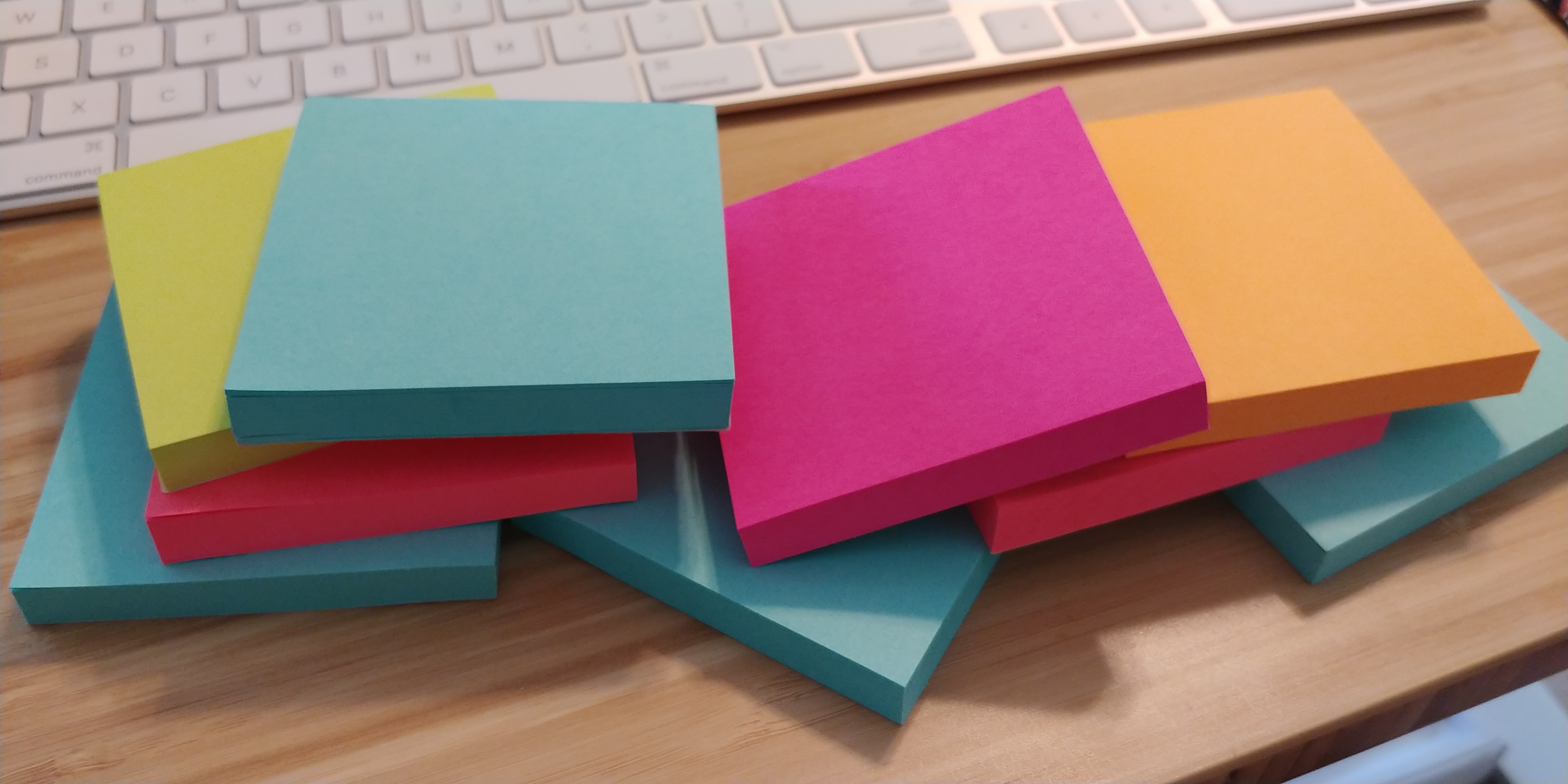 multiple colors of 3x3 sticky note pads, arranged in front off a keyboard in a toppling pile