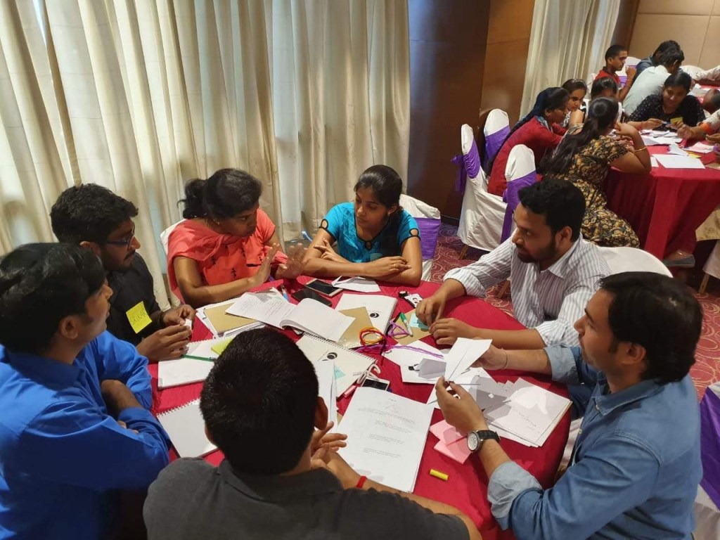 7 people in conversation, sitting at a round table covered in a red cloth with scattered office supplies